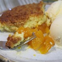 A plate with a piece of the peach cobbler cake, two scoops of ice cream and a bite of the cobbler cake on a fork, ready to eat.