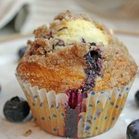One single blueberry muffin on a plate with fresh blueberries surrounding it.