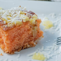 Close up shot of a serving of the fruit punch poke cake with pineapple and coconut sprinkled on the plate and a fork propped on the side of the cake.