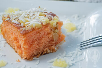 Close up shot of a serving of the fruit punch poke cake with pineapple and coconut sprinkled on the plate and a fork propped on the side of the cake.