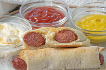 Two halves of a pig in a blanket propped up on a whole one, with condiments in the background.