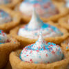 Close up of a sugar cookie cup filled with white frosting and red and blue sprinkles on top.